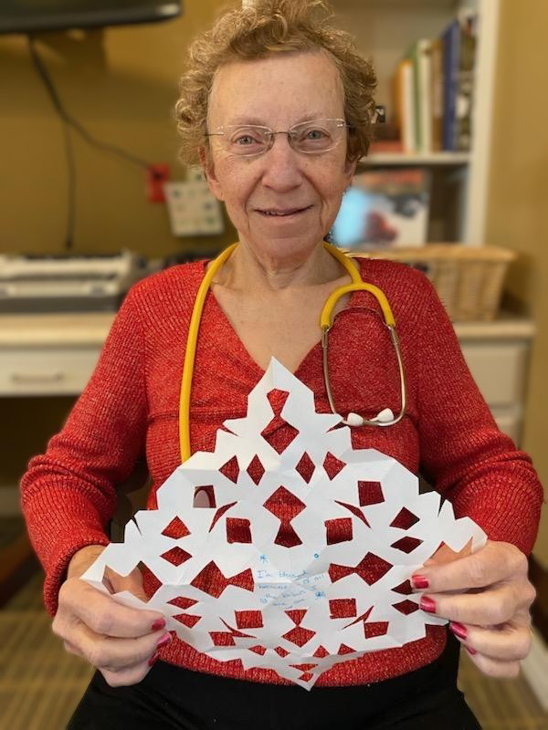 In a recent photo of Mary Ann in a red sweater. She has a yellow stethoscope around her neck and holding a paper snowflake.