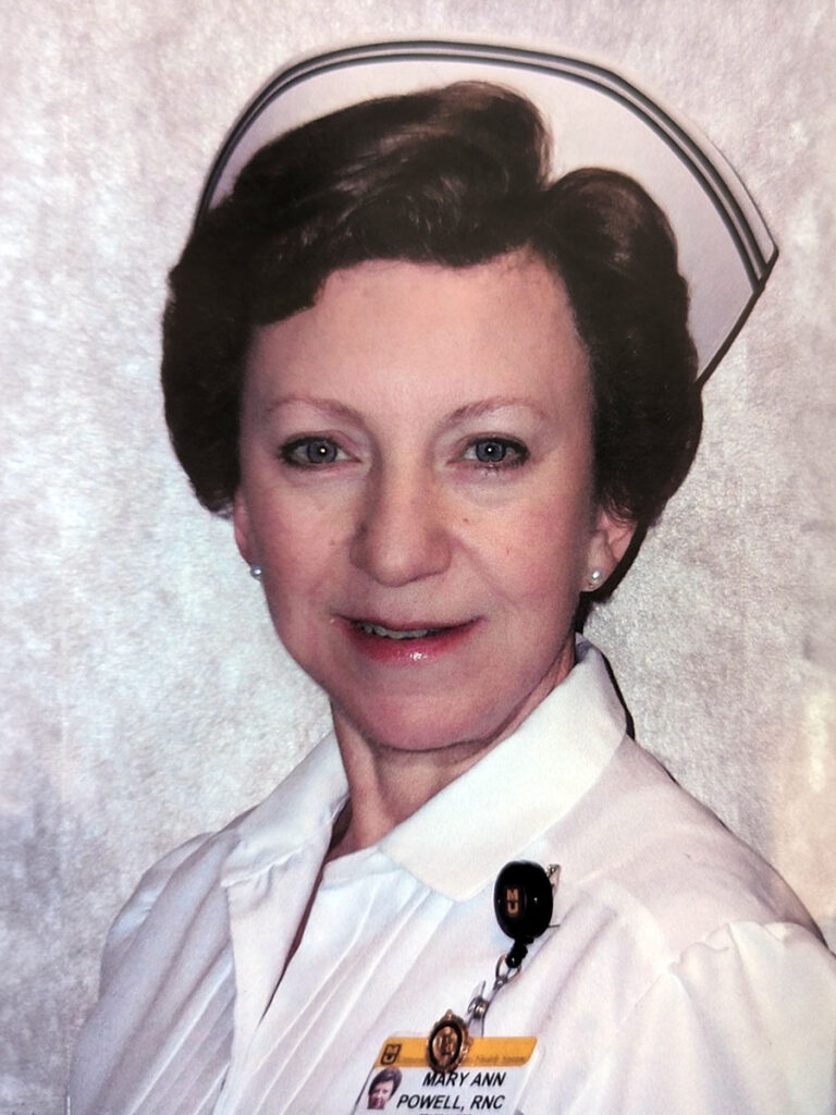 This is a past photo of Mary Ann Powell in her Registered Nurse all-white outfit. Her hat has two lines, and her name badge is clipped to her collared shirt.