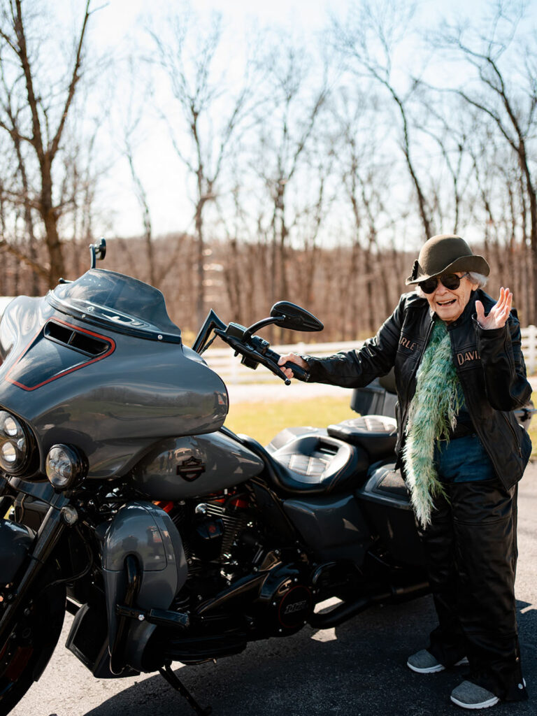 Celebrating Virginia's 100th Birthday with a photoshoot, a woman strikes a pose next to a sleek motorcycle, radiating elegance and excitement.