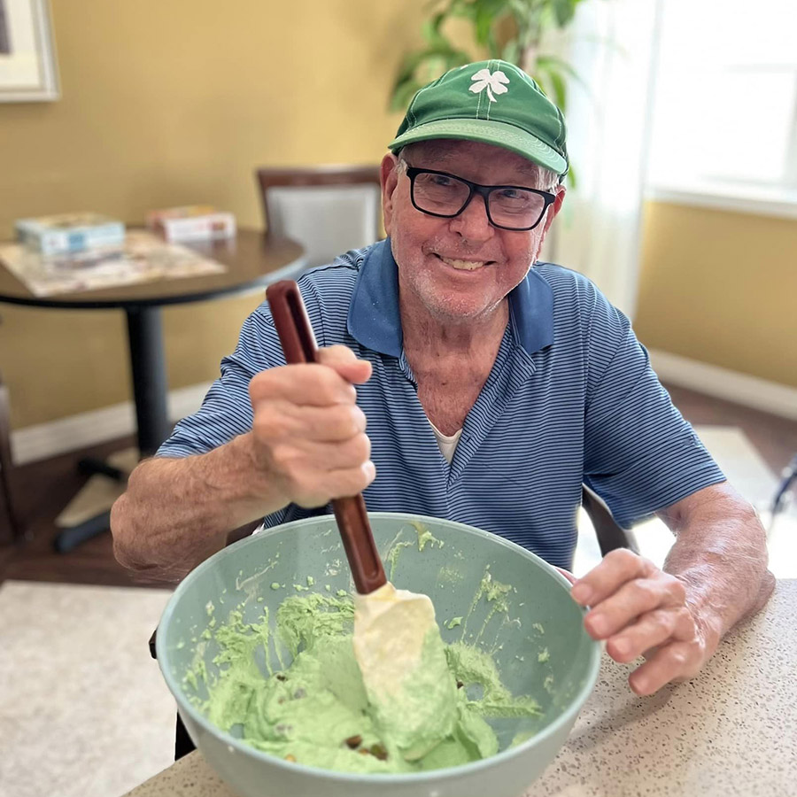 A joyful senior resident stirs homemade ice cream with green food coloring, celebrating St. Patrick's Day with a smile.