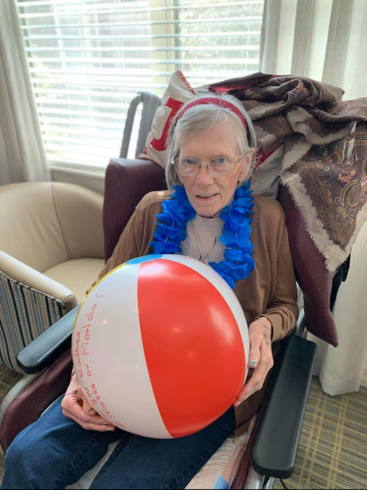 Elderly woman wearing a blue lei and holding a beach ball while sitting in a chair at The Boulevard Wentzville Senior Living.