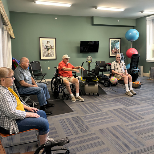 Five senior women sitting in a fitness room, participating in a seated exercise activity with a colorful ball at a senior community.