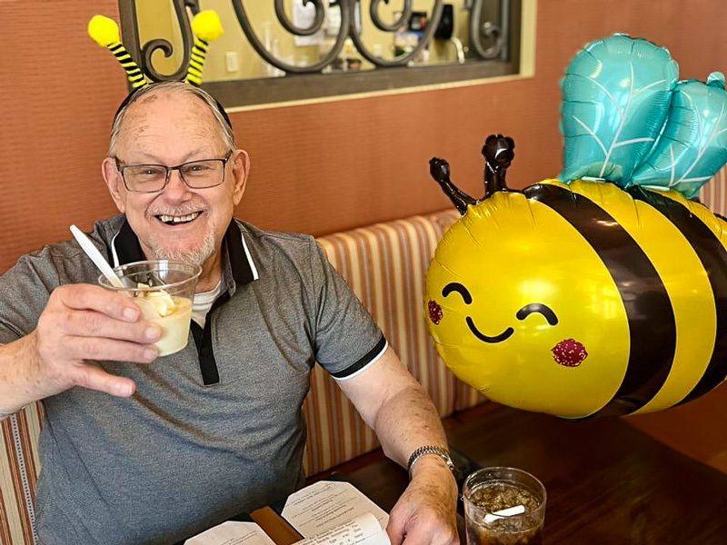 Senior resident smiling and wearing bee antennae headband while holding a dessert cup, sitting next to a cheerful bee balloon in a cozy dining setting.