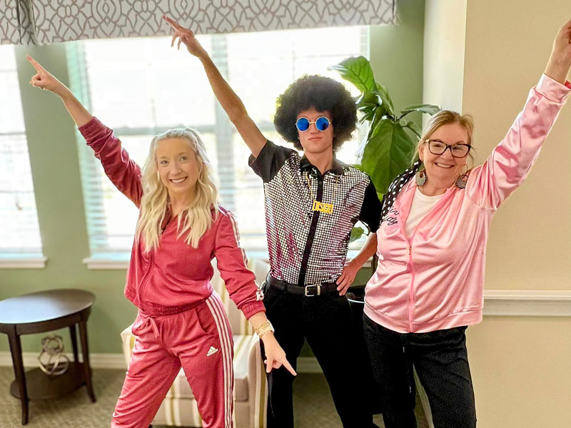 Three senior living team members dressed in 70s-themed attire, striking disco poses with enthusiasm and colorful retro outfits in a brightly lit room.