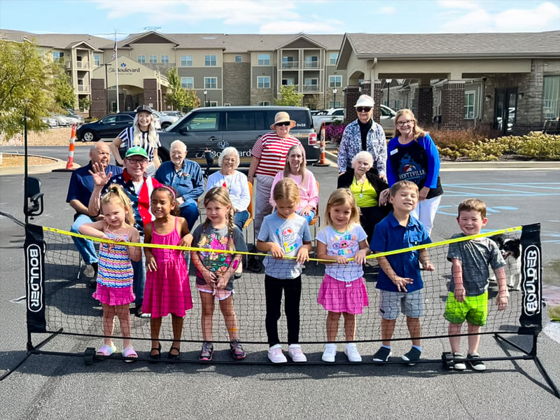 Group photo of senior residents and young children enjoying a sunny outdoor activity, standing behind a volleyball net in the parking area of a senior living community.