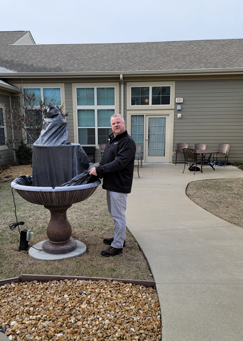 Man stands next to a covered outdoor water fountain in a senior living courtyard, preparing for spring with patio tables and chairs nearby.