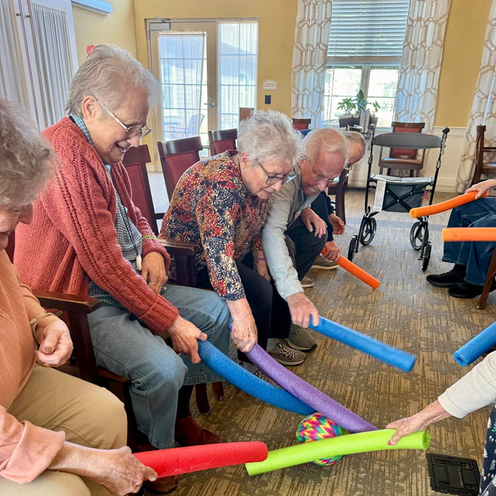 Senior residents participate in a group wellness game using colorful foam noodles to tap a ball, encouraging movement, teamwork, and plenty of smiles.