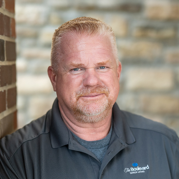 Robert Farmer, Plant Operations Director at The Boulevard Senior Living of Wentzville, smiling while leaning against a brick wall in a dark gray polo shirt with the community logo.