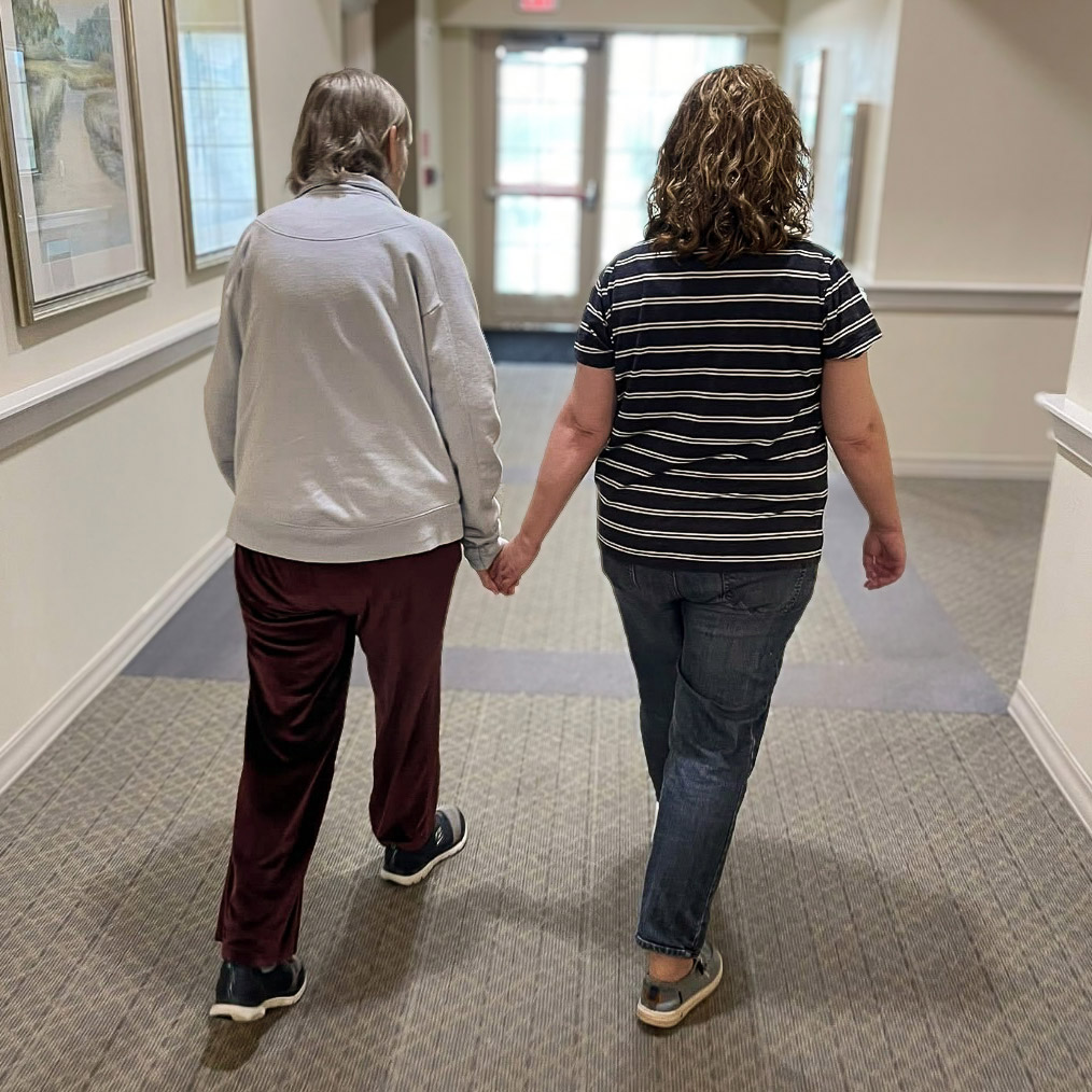 A resident and team member walk hand in hand down a bright hallway, showing care, support, and companionship in the welcoming senior living community.