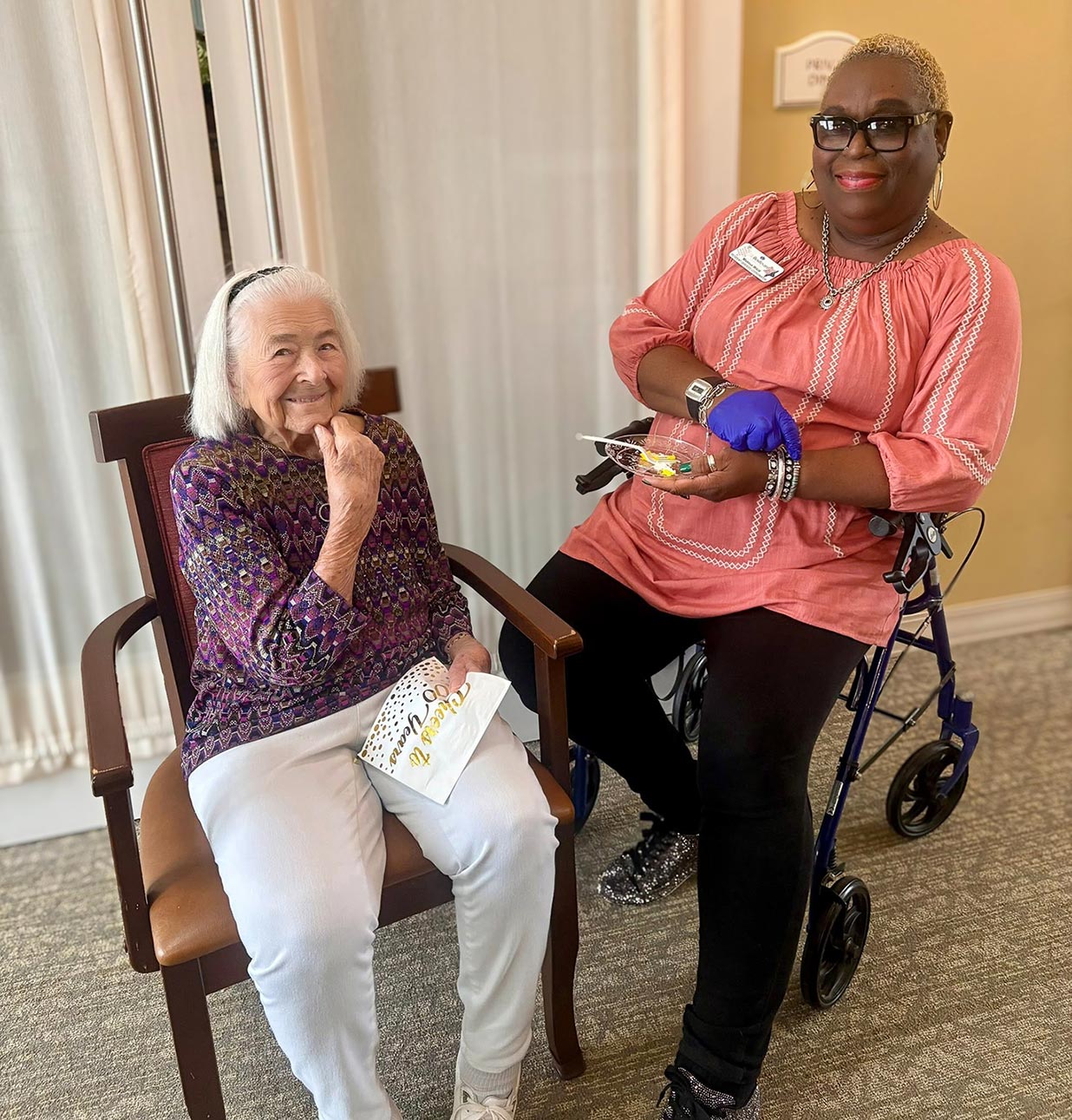 A senior resident and a team member share a cheerful moment indoors, enjoying a sweet treat together. The team member holds a dessert while the resident smiles warmly from her chair.