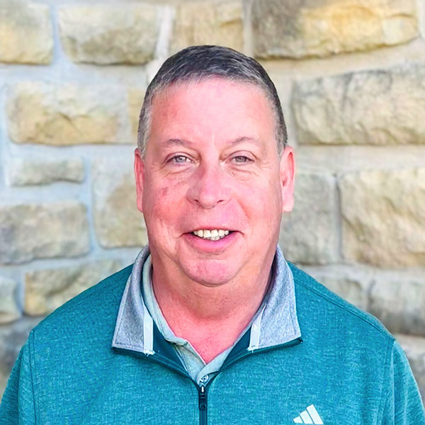 Brian Manhardt, Culinary Director at The Boulevard Senior Living of Wentzville, smiling in a professional headshot, wearing a teal quarter-zip pullover, photographed against a light stone wall background.