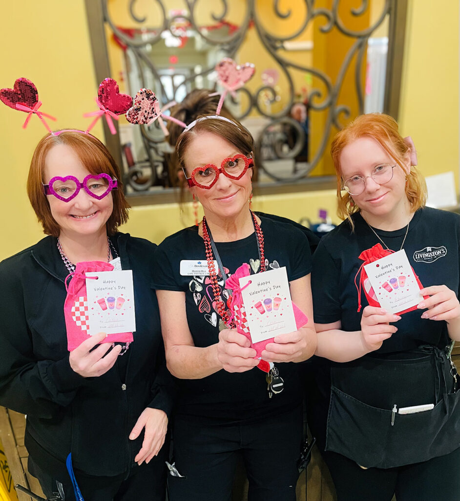 Three team members at Boulevard Wentzville smile, wearing Valentine's props and holding gift bags.
