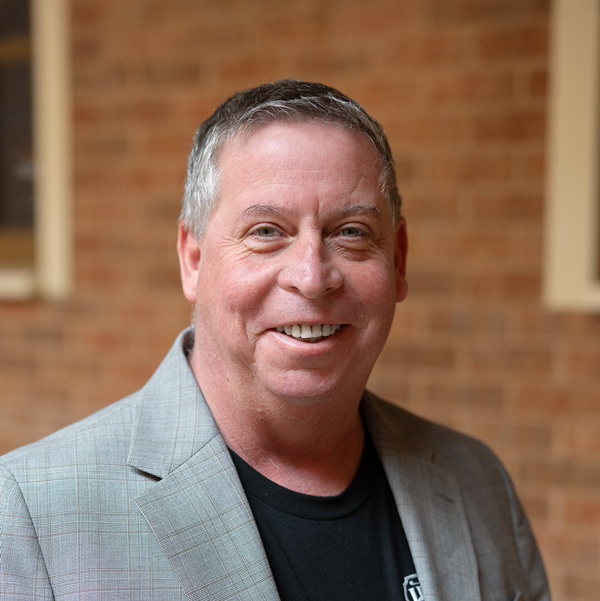 Brian Manhardt, Culinary Director at The Boulevard Senior Living of Wentzville, smiling in a professional headshot, wearing a gray blazer over a black shirt, with a softly blurred brick and window background.