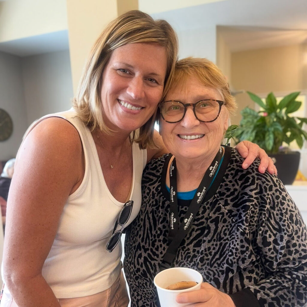 A senior woman holding a cup of coffee smiles alongside a younger woman at The Boulevard Senior Living of Wentzville.