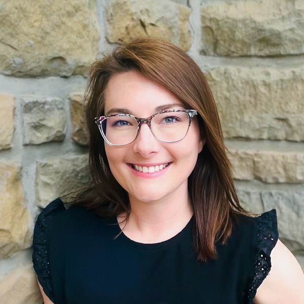 Callee Steffler, Senior Living Coordinator at The Boulevard Senior Living of Wentzville, smiling in a professional headshot with auburn hair, patterned glasses, a black blouse, and a light stone wall background.