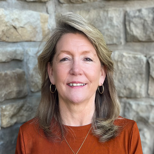 Mary Jane Harris, Executive Director at The Boulevard Senior Living of Wentzville, looking at the camera in a professional headshot with shoulder-length light brown hair, hoop earrings, a rust-colored top, and a light stone wall background.