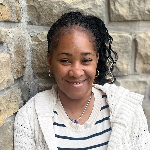 Beverly Warren, Memory Care Director at The Boulevard Senior Living of Wentzville, smiling in a professional headshot with braided hair pulled back, hoop earrings, a striped top, a light knit cardigan, and a light stone wall background.