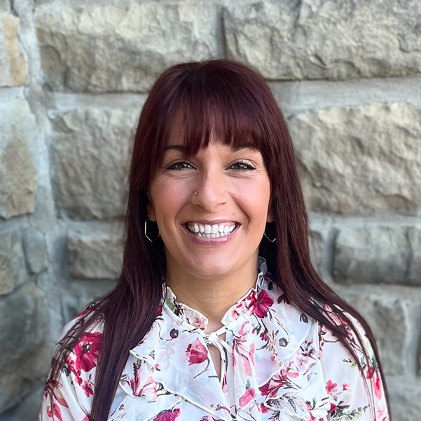 Carissa Stogsdill, Operations Coordinator at The Boulevard Senior Living at Wentzville, smiling in a professional headshot with long dark auburn hair, a floral blouse, and a light stone wall background.