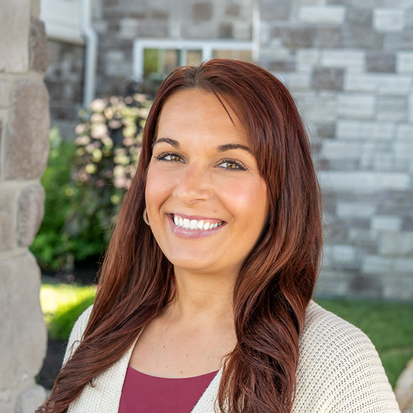 Carissa Stogsdill, Operations Coordinator at The Boulevard Senior Living of Wentzville, smiling in a professional outdoor headshot with long auburn hair, a light cardigan, and a softly blurred stone building background.