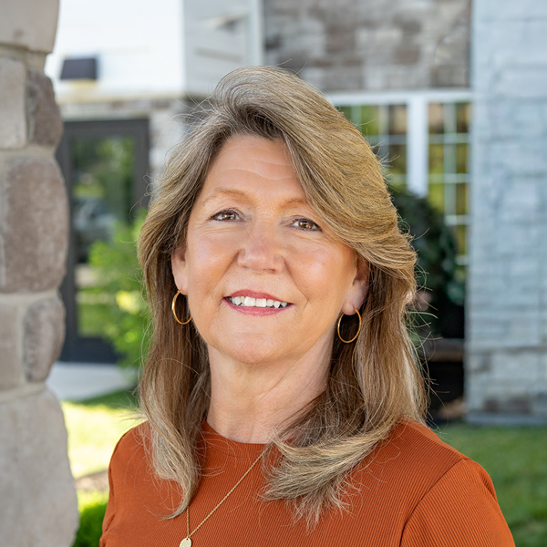 Mary Jane Harris, Executive Director at The Boulevard Senior Living of Wentzville, looking at the camera in a professional outdoor headshot with light brown hair, gold hoop earrings, a rust-colored top, and a softly blurred stone building background.