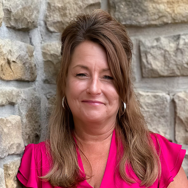 Jodie Enger, Wellness Director at The Boulevard Senior Living of Wentzville, looking at the camera in a professional headshot with long light brown hair, silver hoop earrings, a bright pink blouse, and a light stone wall background.